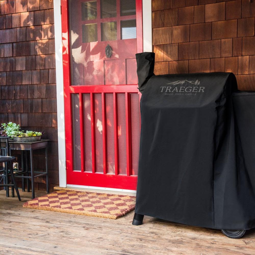 Black full-length grill cover with Traeger logo protecting outdoor grill in rustic porch setting for grilling accessories