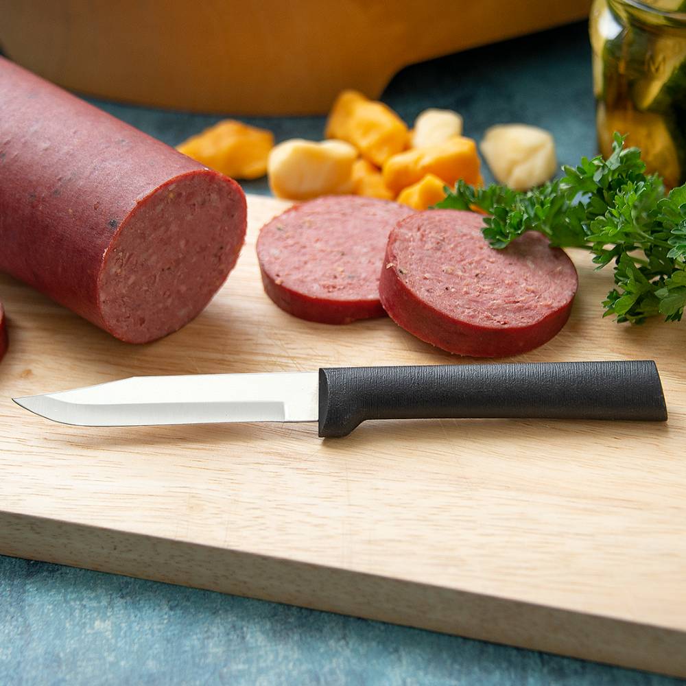 Paring knife with black handle and sharp stainless steel blade on cutting board with sliced sausage and herbs
