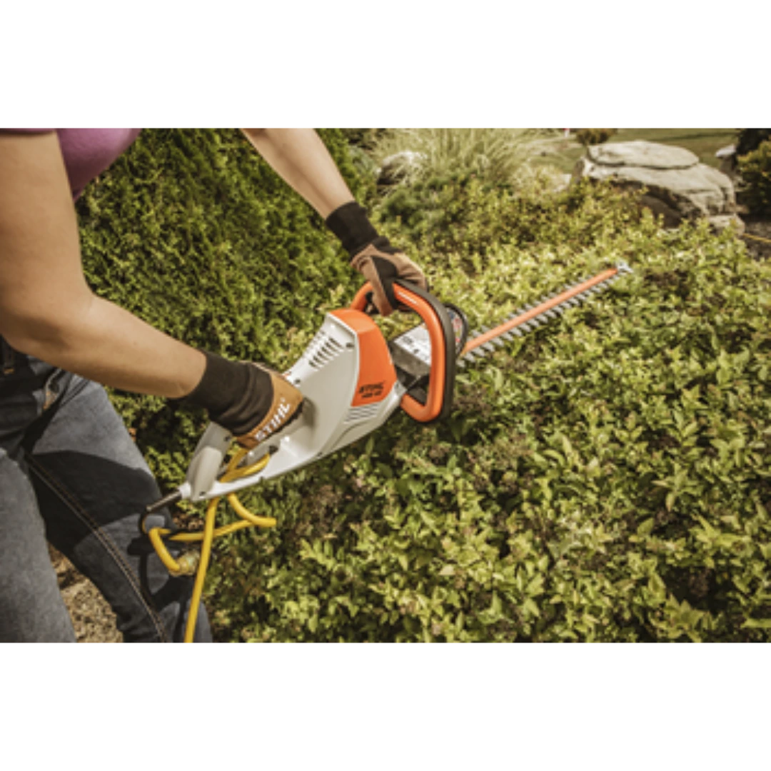 Person wearing gloves using electric hedge trimmer to shape dense green shrubbery for home improvement tasks