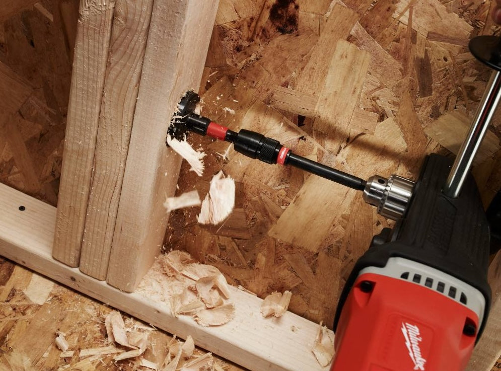 Red and black Milwaukee selfeed drill bit attached to power drill cutting large hole in wood framing lumber