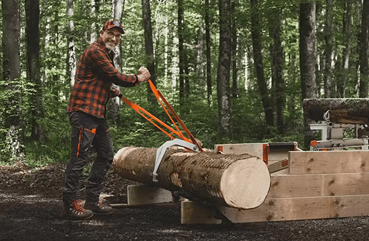 Man using a LOGOSOL Smart Turner XL with orange handles to maneuver a large log in a forest setting for woodworking tasks