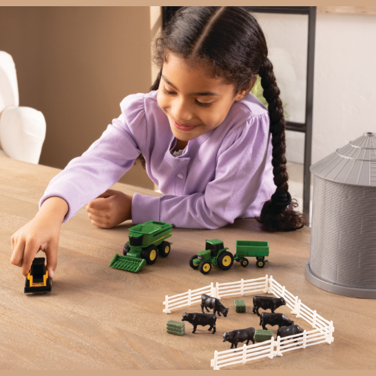 Child playing with green toy tractor, black farm animals in white fence, and gray grain bin on wooden table