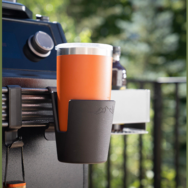 Black plastic cup holder with mountain design securely attached to grill for outdoor grilling and lawn accessories.