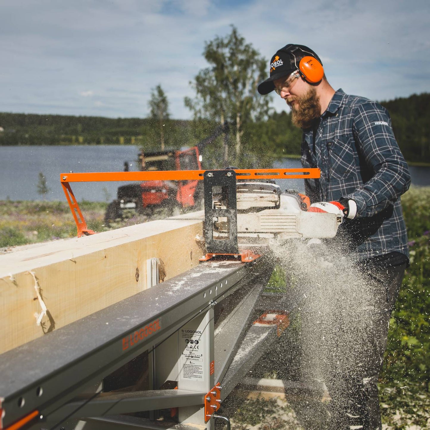 Man wearing protective gear using a chainsaw with orange steering bar to cut large timber outdoors near water