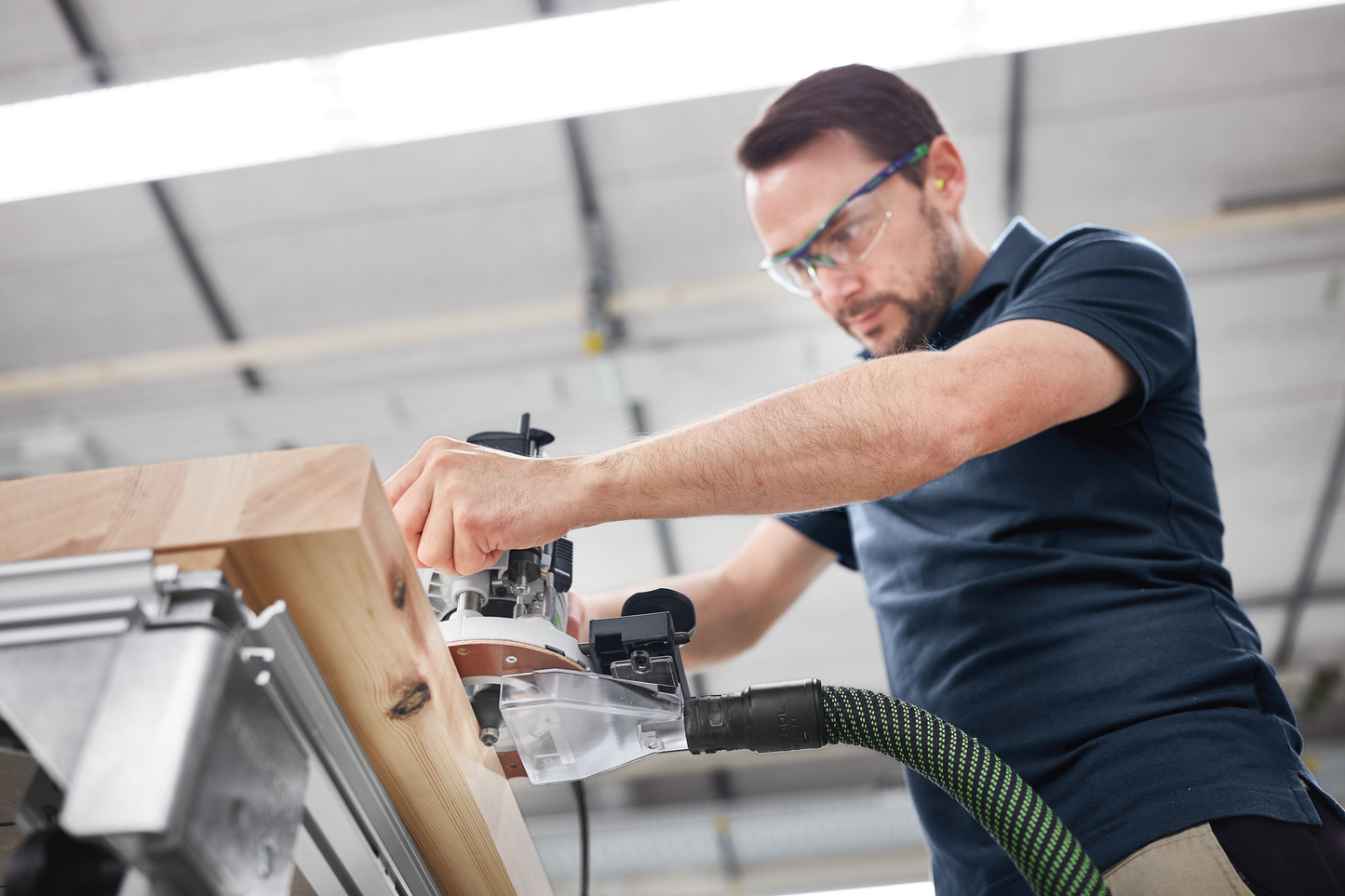 Worker wearing safety glasses using a woodworking router on thick wood in a bright workshop for home improvement tools