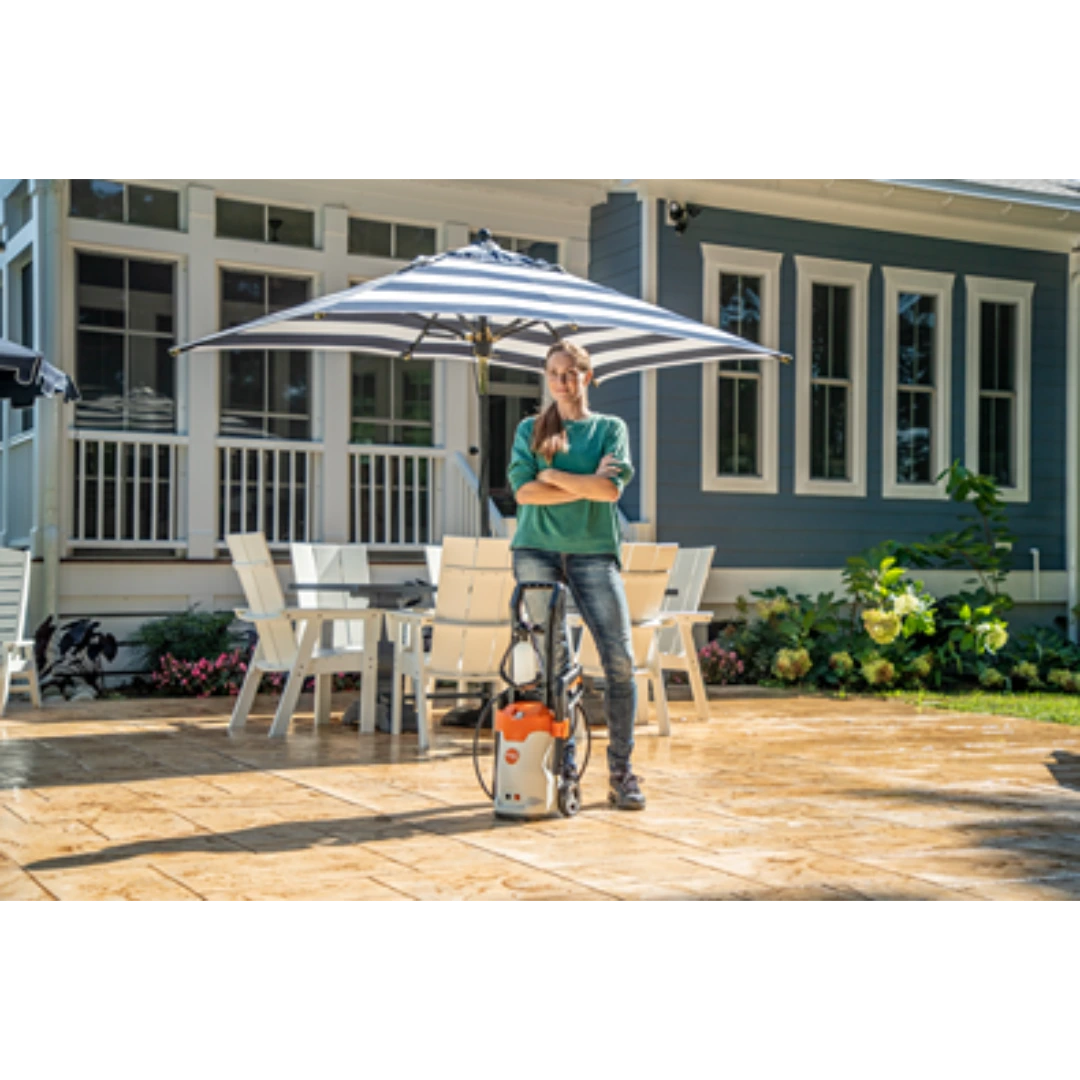 Woman standing outdoors on clean patio holding STIHL electric pressure washer near white patio set and blue house