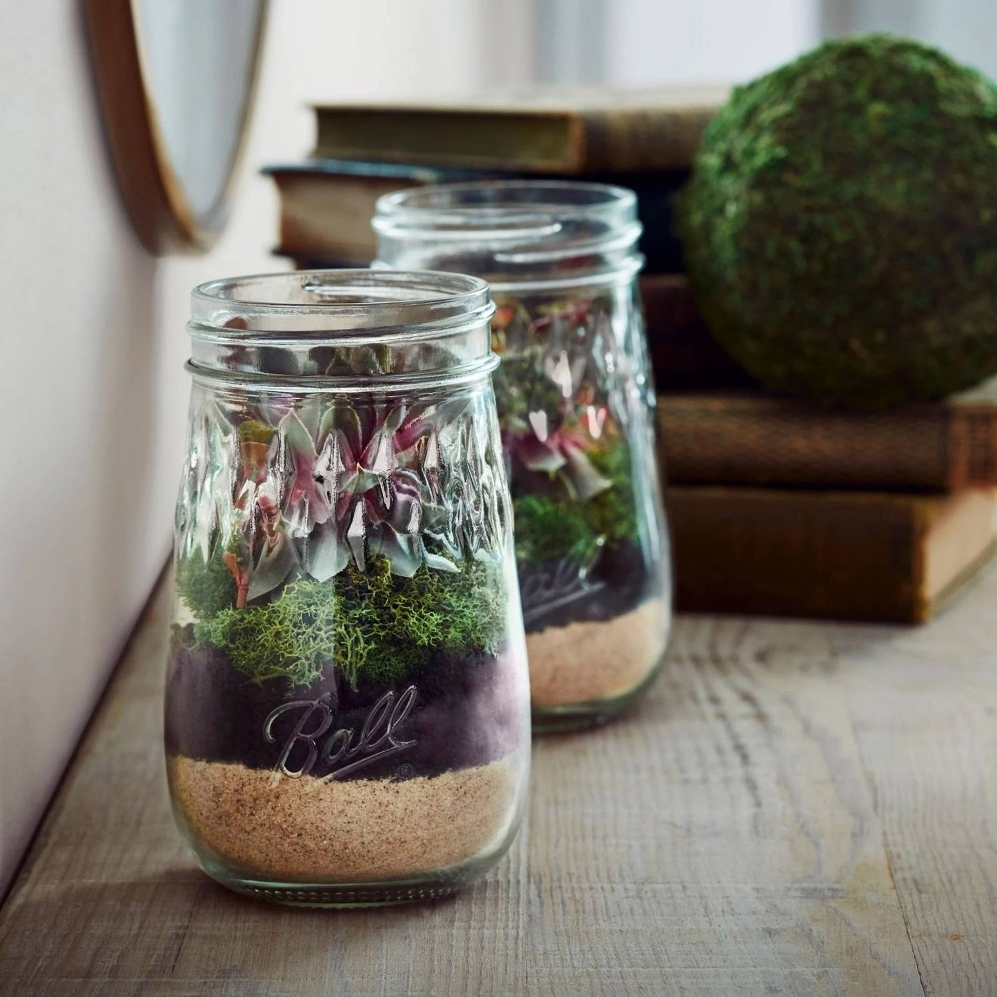 Two clear pint glass jars with layered sand, soil, moss, and decorative flowers on wooden surface in Kitchenware collection