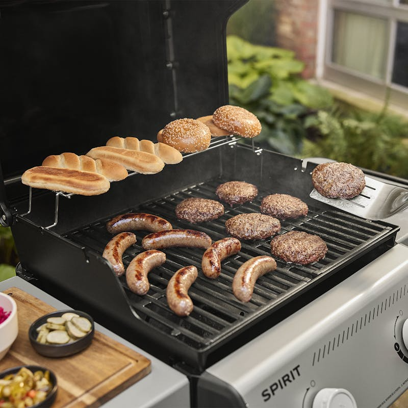 Black propane grill with two burners cooking sausages, burgers, and toasting buns in outdoor living setting