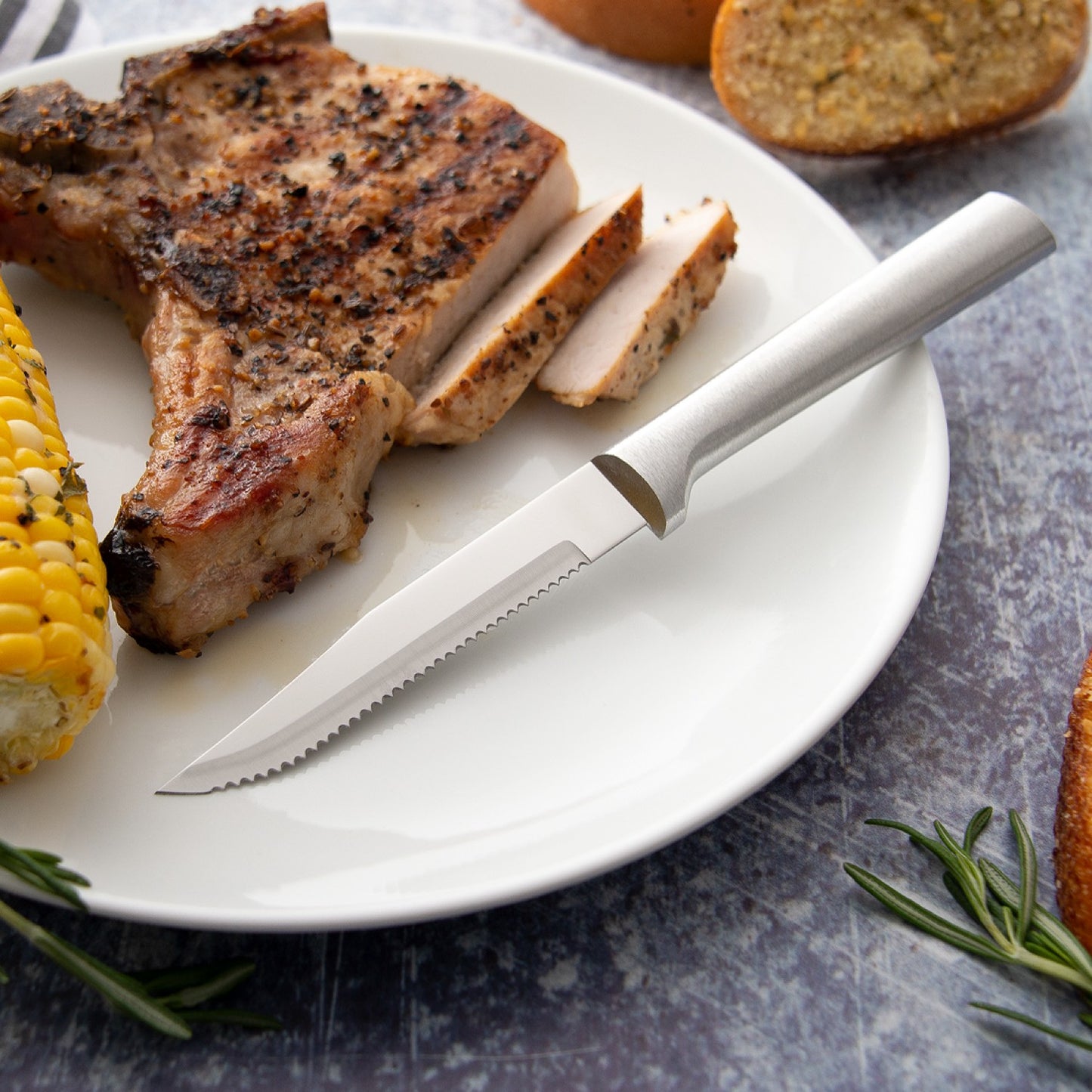 Serrated steak knife with solid aluminum handle on white plate with sliced grilled pork chop, corn on the cob, and fresh rosemary, ideal for Kitchenware and Cooking Utensils under $50.