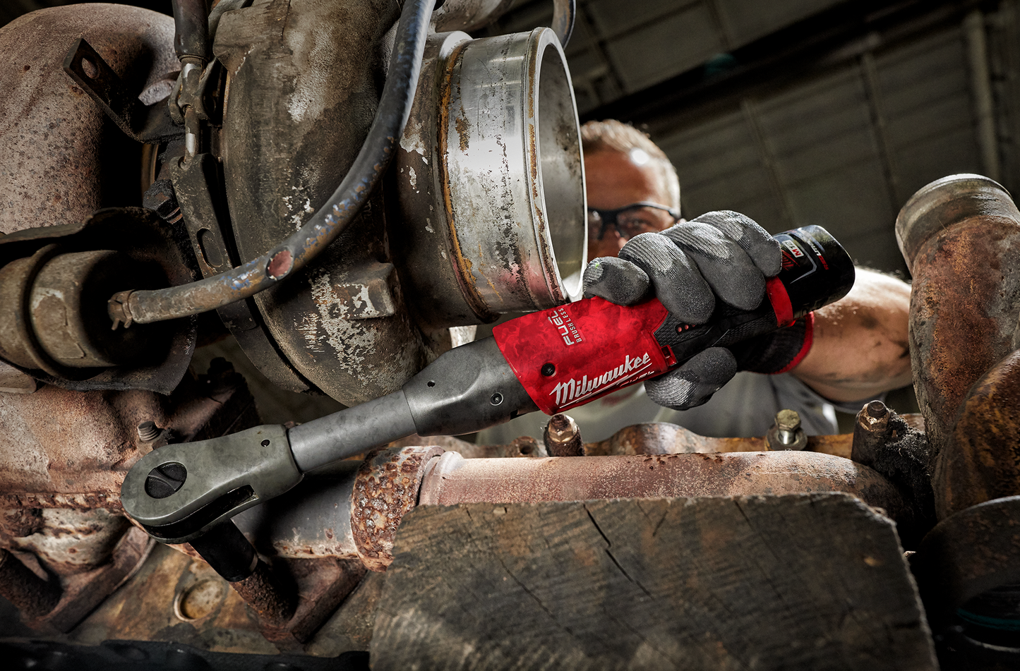 Gloved hand using red Milwaukee cordless extended reach ratchet on heavy machinery for repair work in workshop