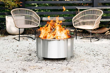 Outdoor stainless steel fire pit with bright flames and modern patio chairs in an outdoor living setting