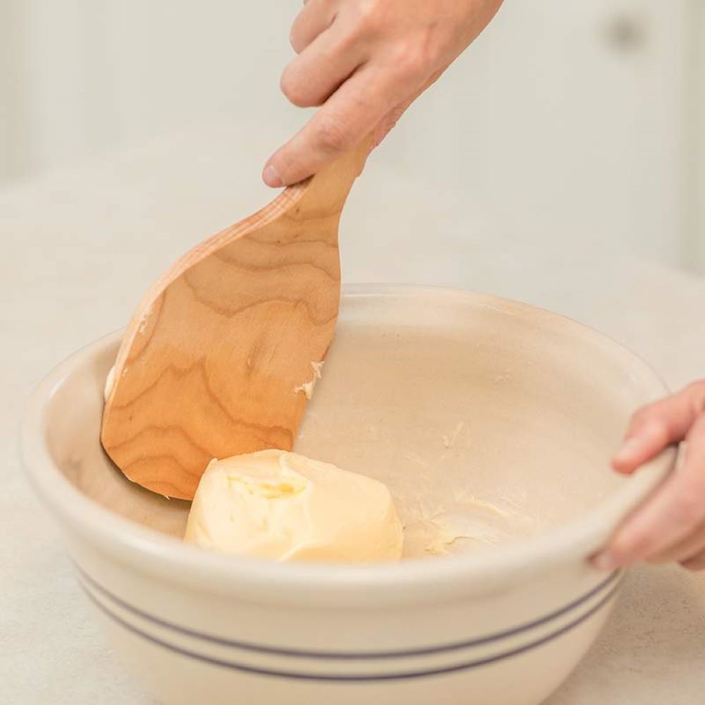 Hand holding a wood paddle spreading softened butter in a ceramic mixing bowl for kitchenware collections
