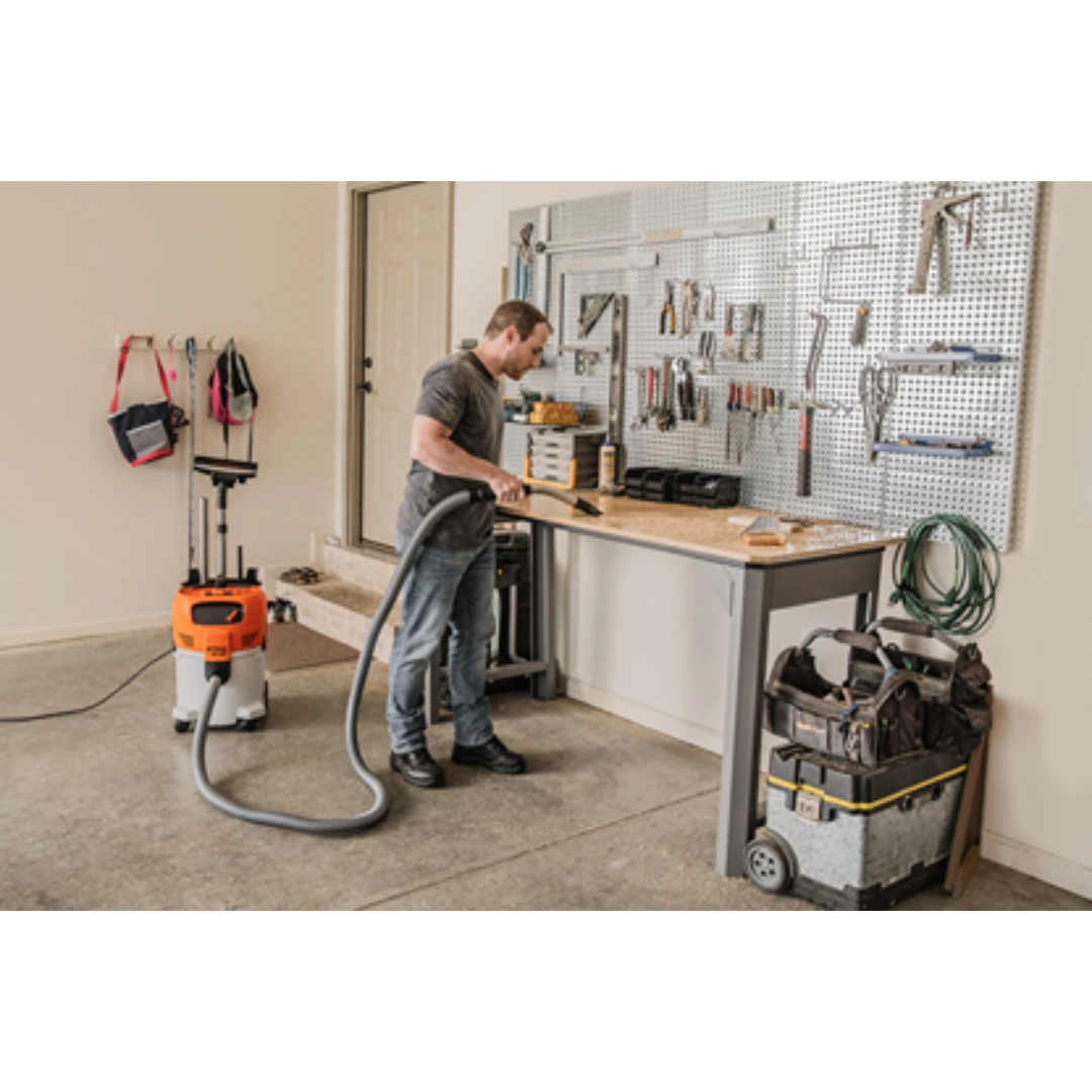 Man using an orange and white wet/dry vacuum with flexible hose to clean a workshop floor near a workbench and tools
