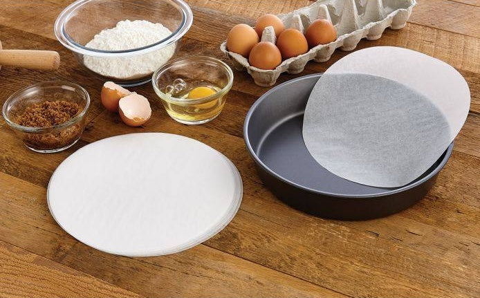 Ten-inch round parchment paper sheets placed next to baking pan with eggs and ingredients on wooden counter