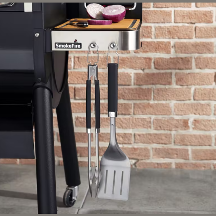 Stainless steel grill tongs and slotted spatula with soft grip handles hanging on smoker side shelf in outdoor living collection