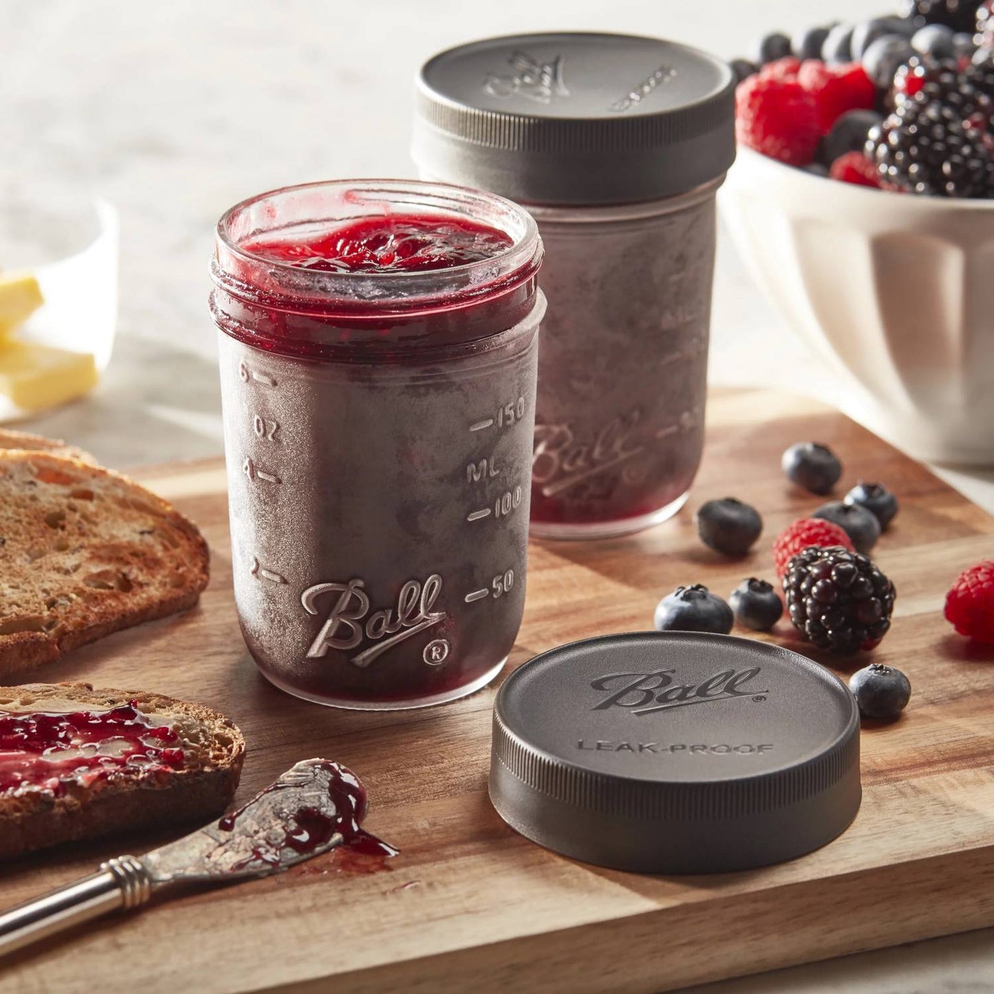 Two glass jars with black leak-resistant lids, one filled with berry jam, surrounded by fresh berries and toast on wooden board