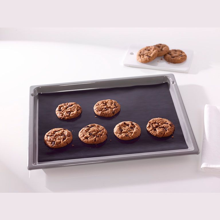 Gray baking sheet lined with reusable nonstick liner holding six chocolate chip cookies in kitchenware collection