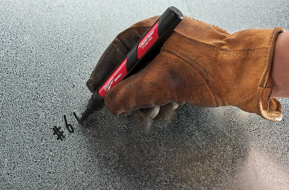 Hand wearing brown work glove using black paint marker to write on rough metallic surface, woodworking hand tool in use