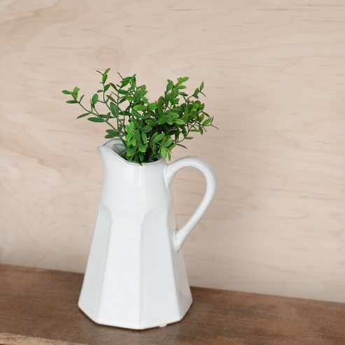 White ceramic decorative vase with geometric facets holding green leafy stems on wooden surface