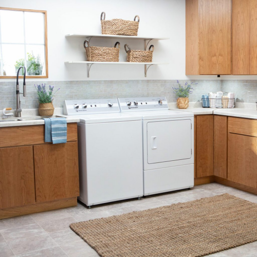 White 26-inch top load washer paired with matching dryer in a bright laundry room with wooden cabinets and woven baskets