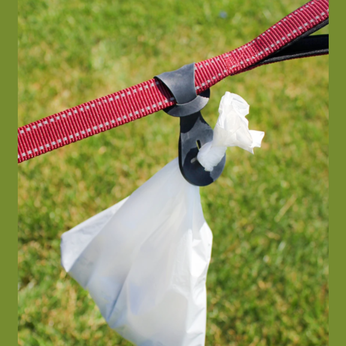 Black plastic pet waste bag holder hanging from a red dog leash outdoors on green grass