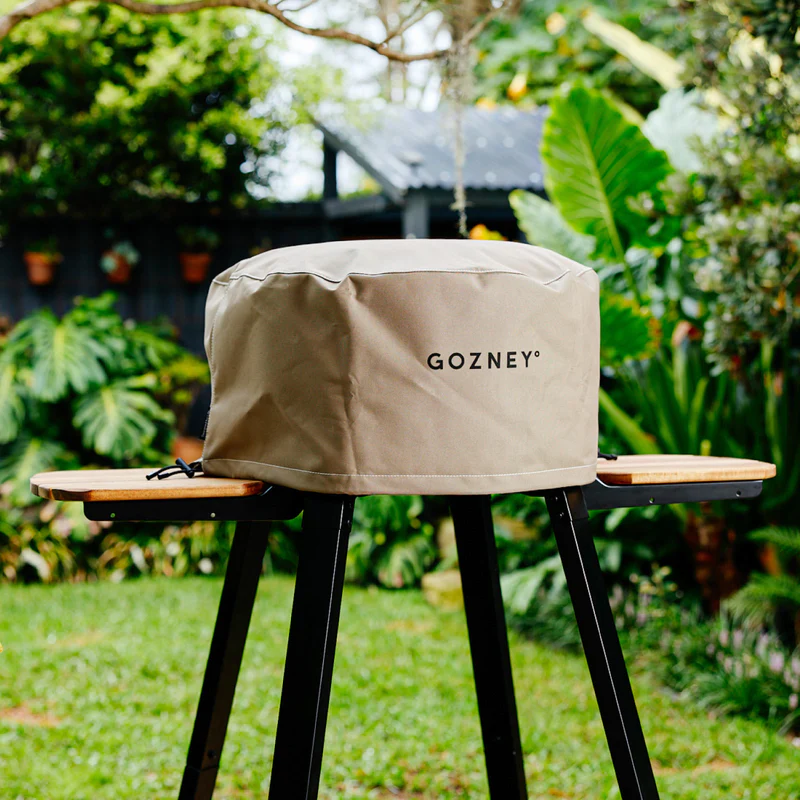 Marine grade weather-resistant tan grill cover with black logo protecting outdoor cooking equipment in garden setting