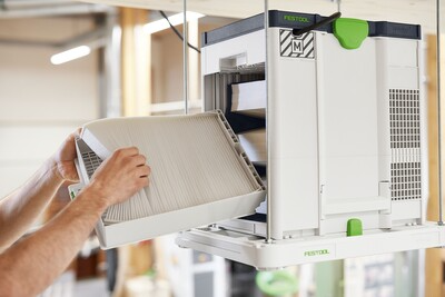 Hands installing a white and green Festool air purifier on a ceiling bracket in woodworking tools and dust filtration setup