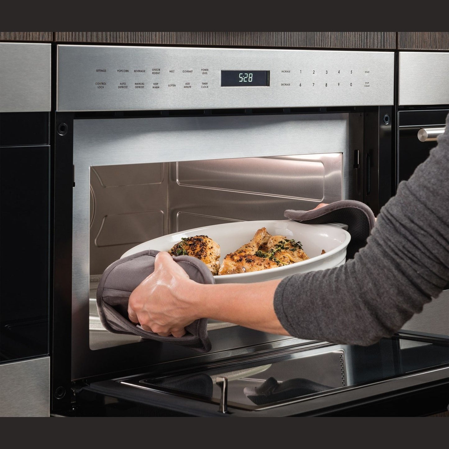 Person using oven mitts to remove roasted chicken from built-in stainless steel drop-down microwave in modern kitchen