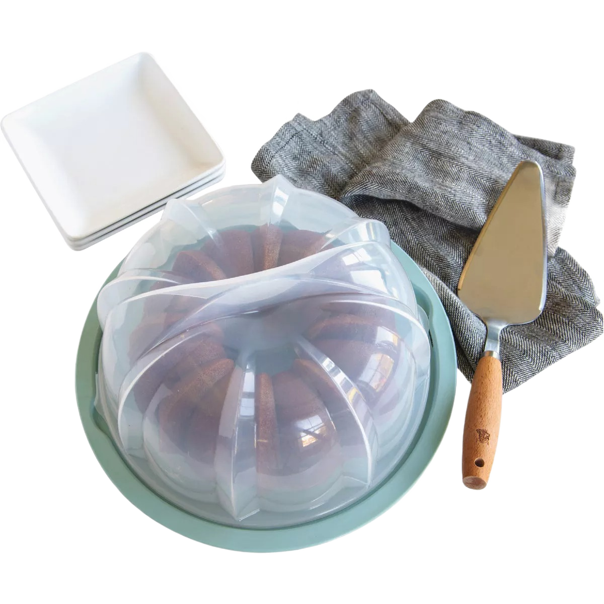Green bundt cake keeper with clear locking dome, gray linen napkin, cake server, and white square plates in kitchen storage setting