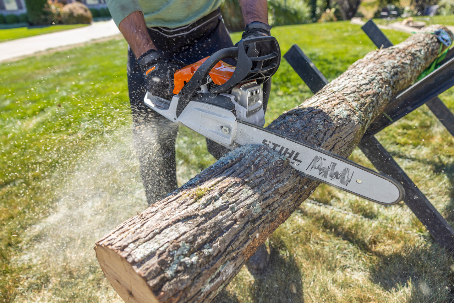 Person cutting a large log outdoors with an orange and white gas-powered chainsaw on a sunny day