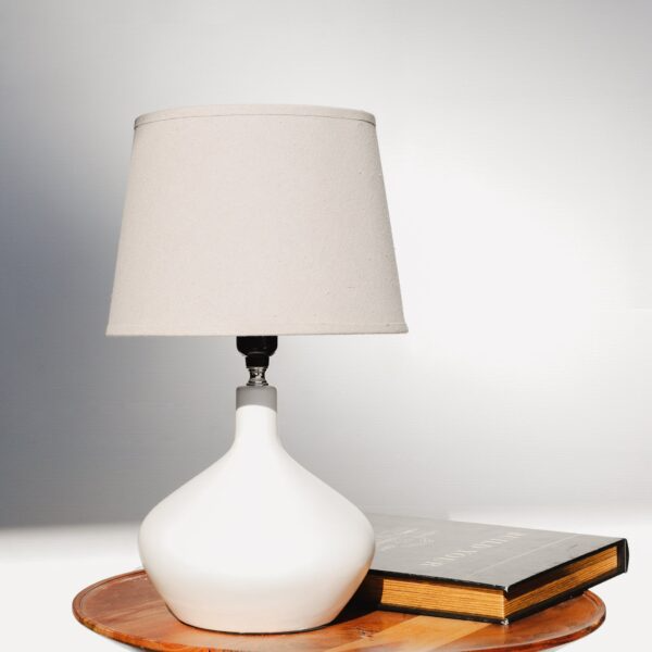 White ceramic table lamp with fabric shade on wooden surface next to a closed hardcover book in lighting collections
