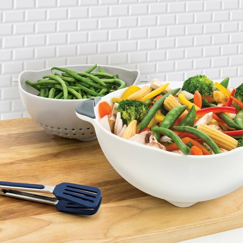 Oyster gray colander with handles holding fresh green beans and a larger bowl with mixed vegetables on wooden counter