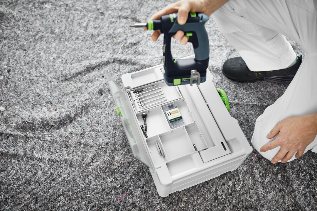 Person holding Festool cordless drill above organized drill bit storage case on textured floor for home improvement