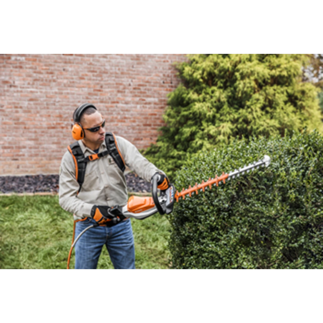 Man wearing protective gear trimming dense hedge with battery-powered orange and white hedge trimmer in a garden