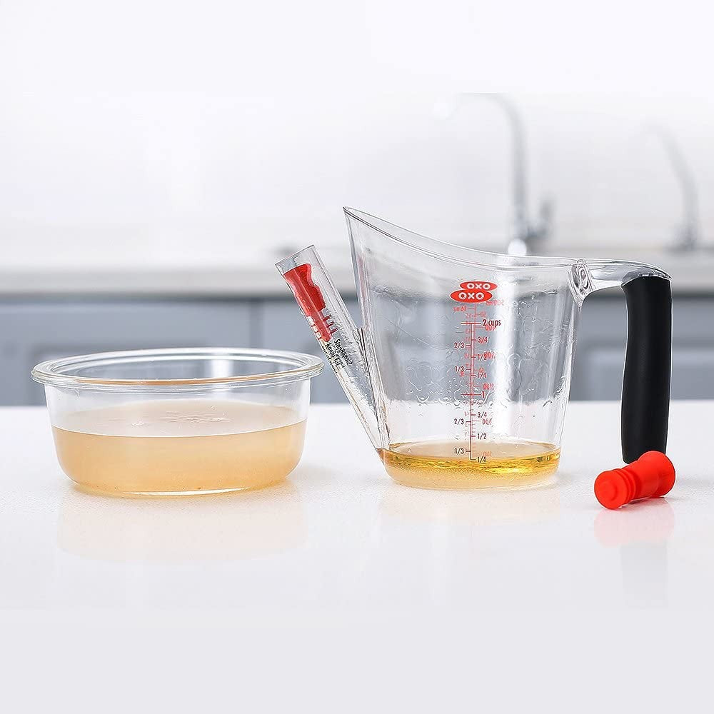 Clear 2 cup fat separator with black handle and red stopper beside bowl of broth in kitchenware and gadgets setting