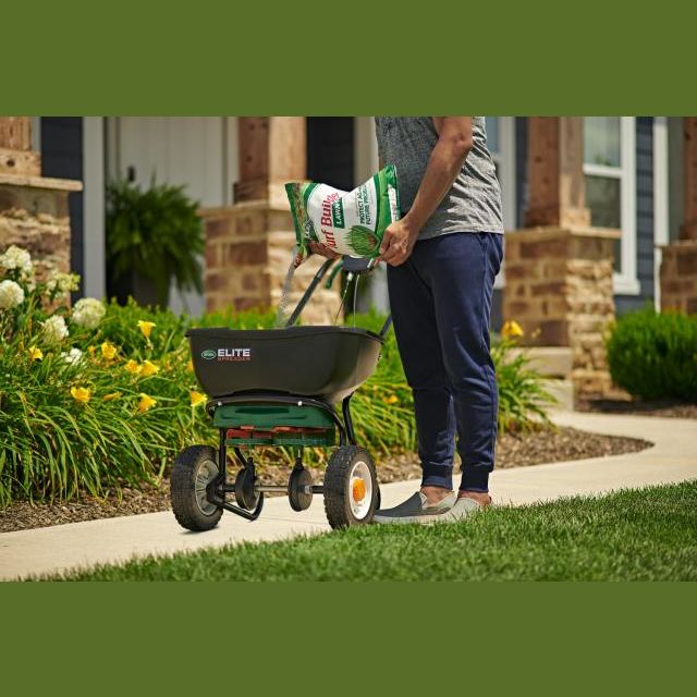 Person pouring Scotts Turf Builder lawn food into a black fertilizer spreader on a garden pathway near a house