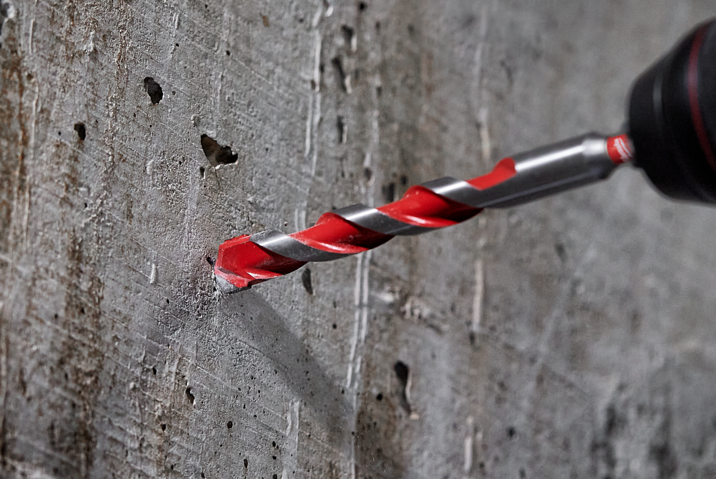 Carbide-tipped hammer drill bit with red spiral flutes drilling into concrete wall in power tool accessories collection