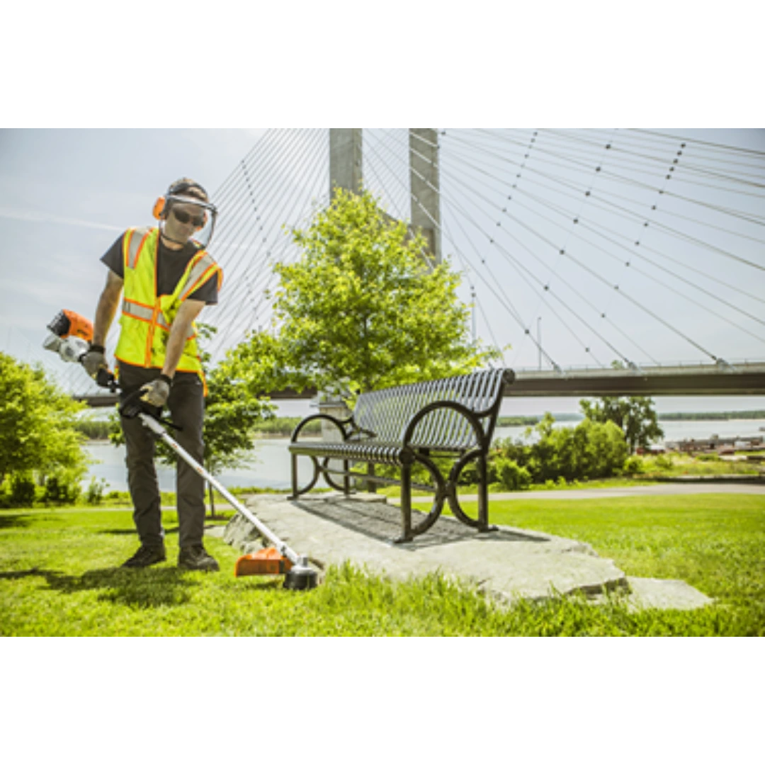 Worker in safety vest using a STIHL KM 91 R KombiMotor to trim grass beside a metal park bench by a bridge