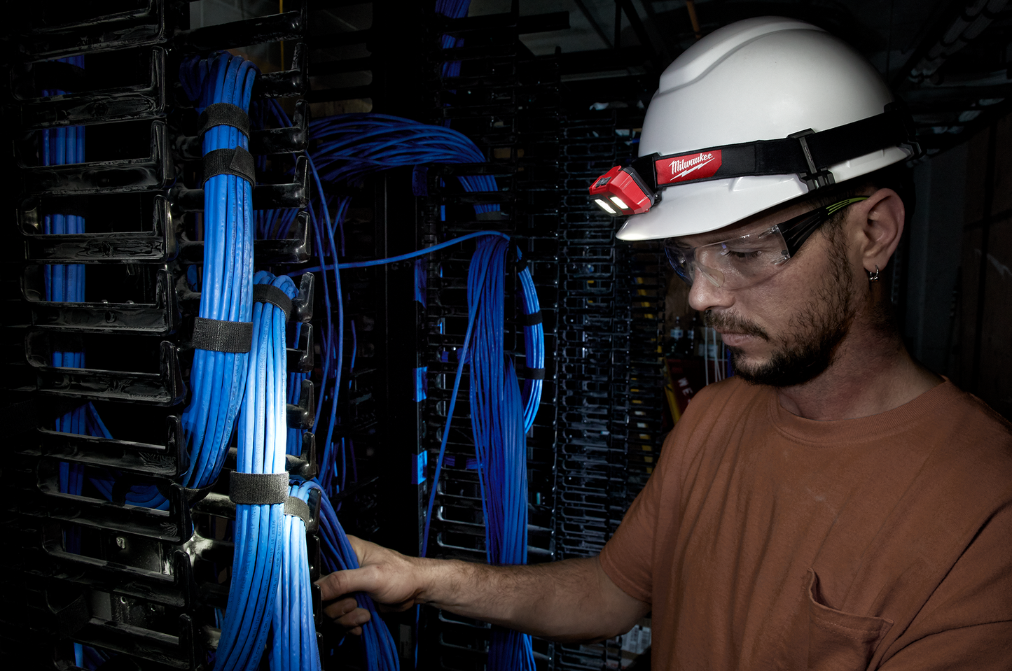 Man wearing Milwaukee headlamp and safety glasses working on organized blue network cables in dark server room