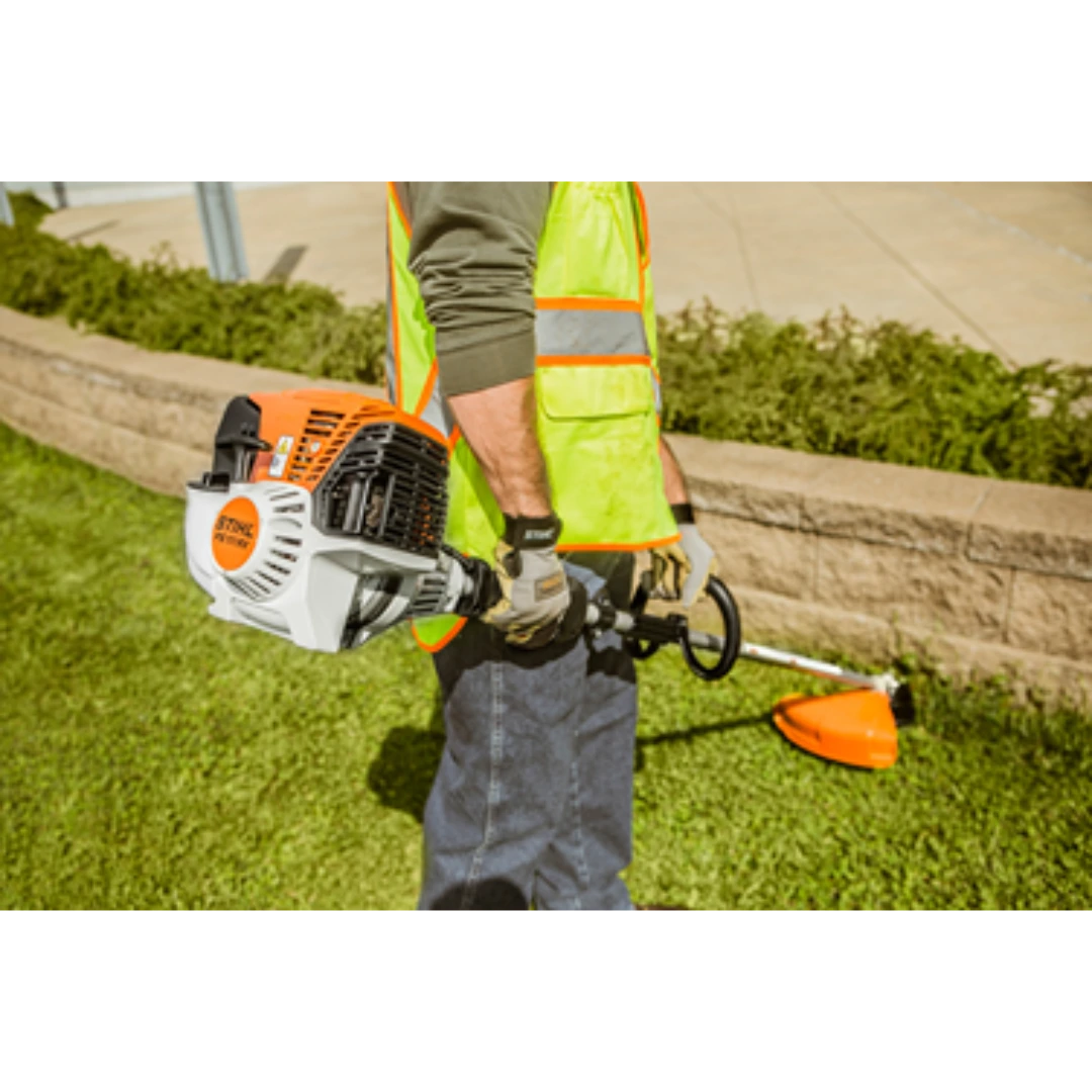 Person wearing safety vest holding a professional straight shaft trimmer with orange and white housing near a retaining wall