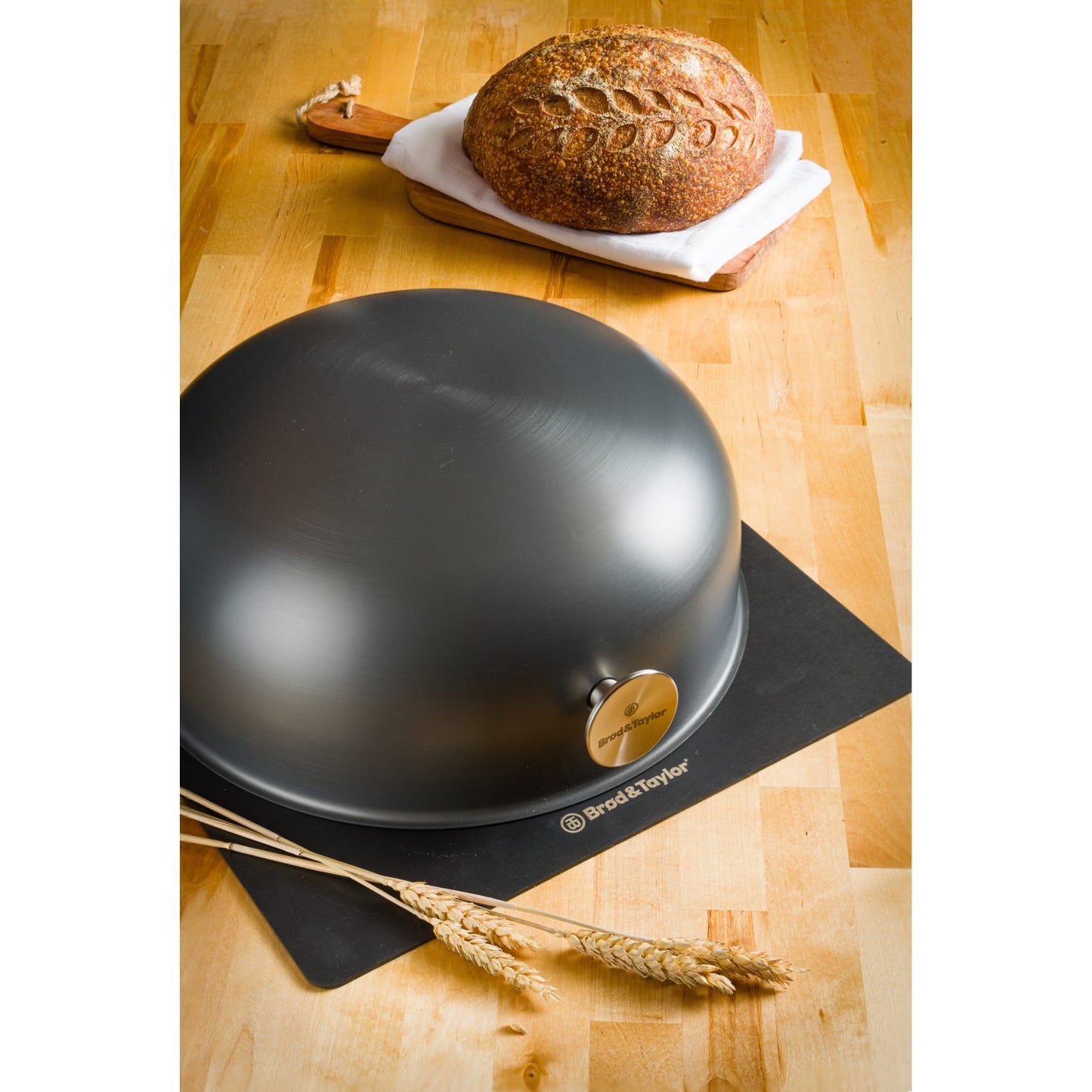 Matte black baking shell dome with branded badge on black silicone mat beside artisan boule and wheat stalks on wood surface