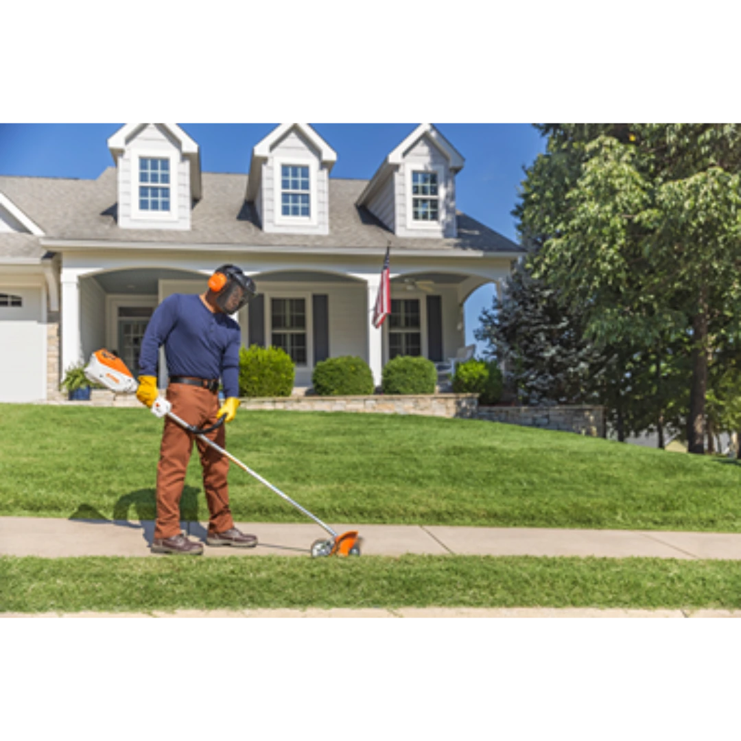 Person wearing protective gear using a battery-powered edger to trim grass along a sidewalk in a suburban yard