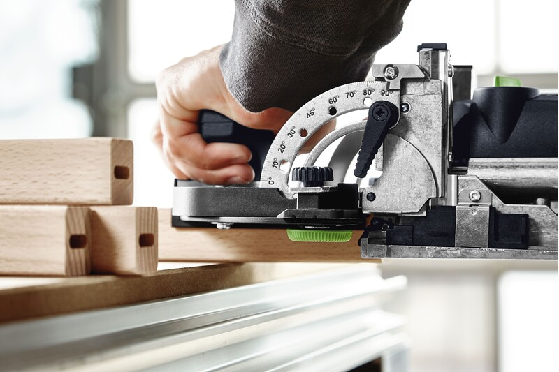 Close-up of a hand operating a Festool woodworking tool with angled adjustment near mortised wooden rails for precise joinery tasks