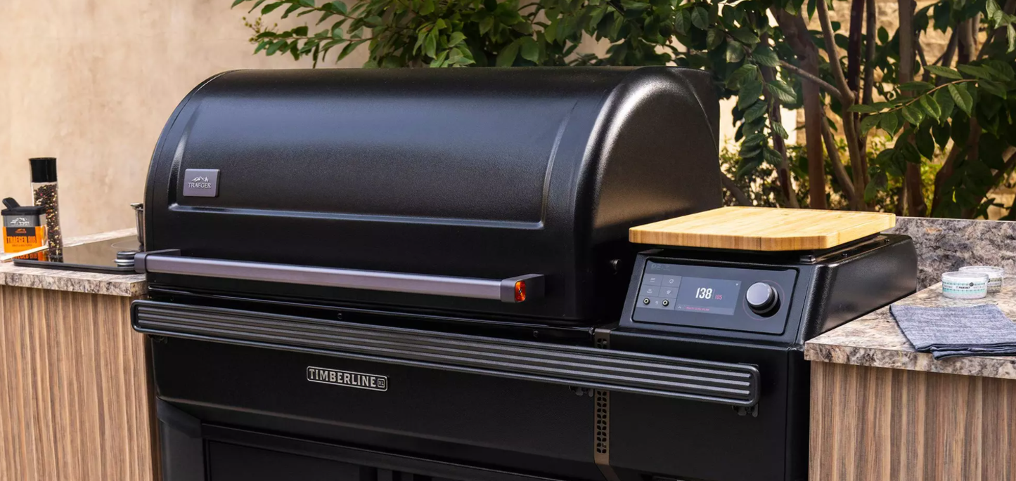 Black pellet grill with digital control panel and wooden cutting board on side shelf in outdoor living space