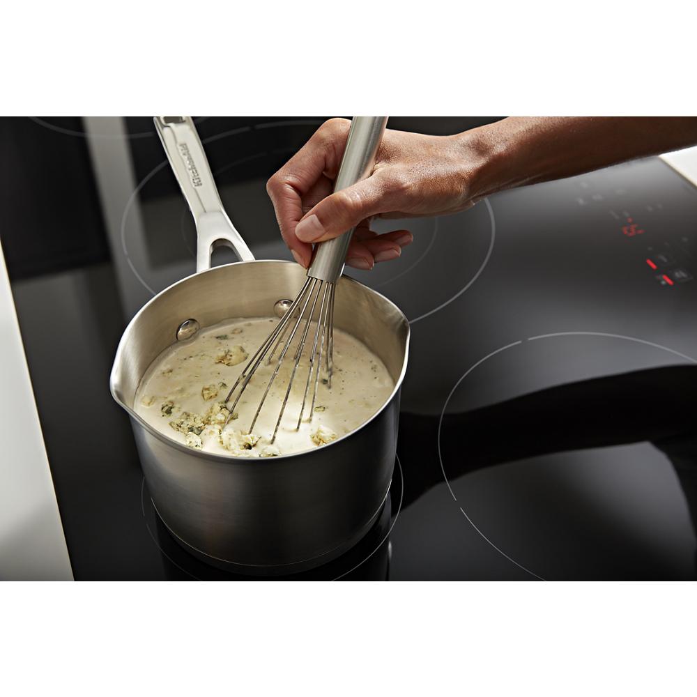 Person whisking sauce in stainless steel saucepan on black sensor induction cooktop with digital controls