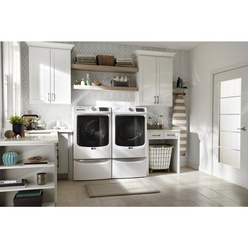 White front load washer and dryer on white storage pedestals in bright laundry room with cabinetry and shelving