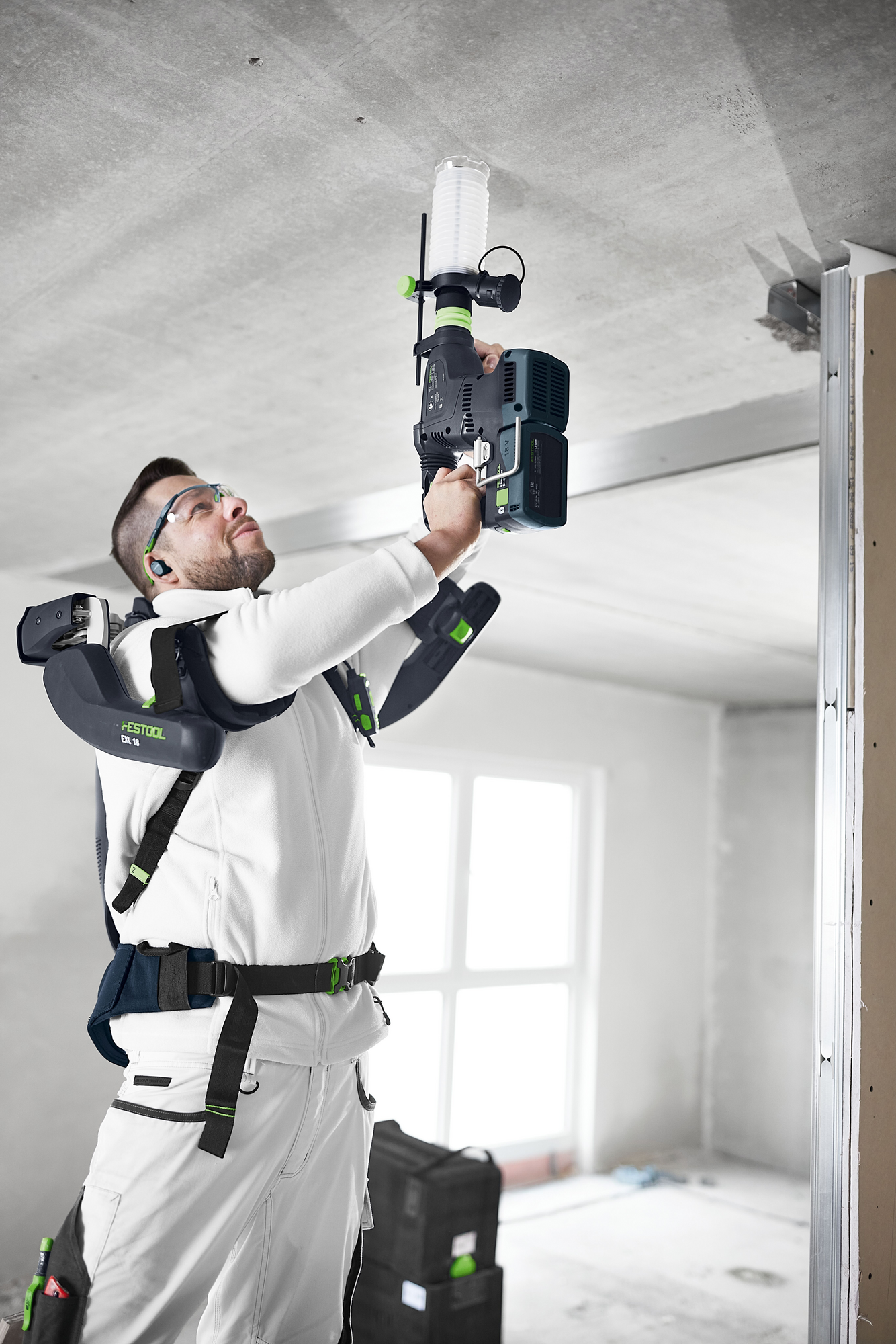 Man wearing ergonomic Festool support device while using cordless drill with auxiliary handle for ceiling work indoors