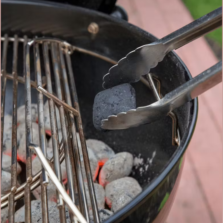 Nickel-plated hinged steel kettle grill grate with charcoal briquettes being added using metal tongs in outdoor grilling setup