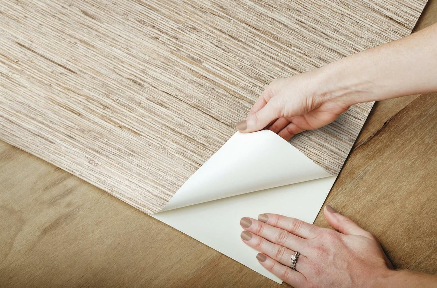 Close-up of hands applying neutral-toned peel and stick wallpaper with faux weave grasscloth texture on a wooden surface