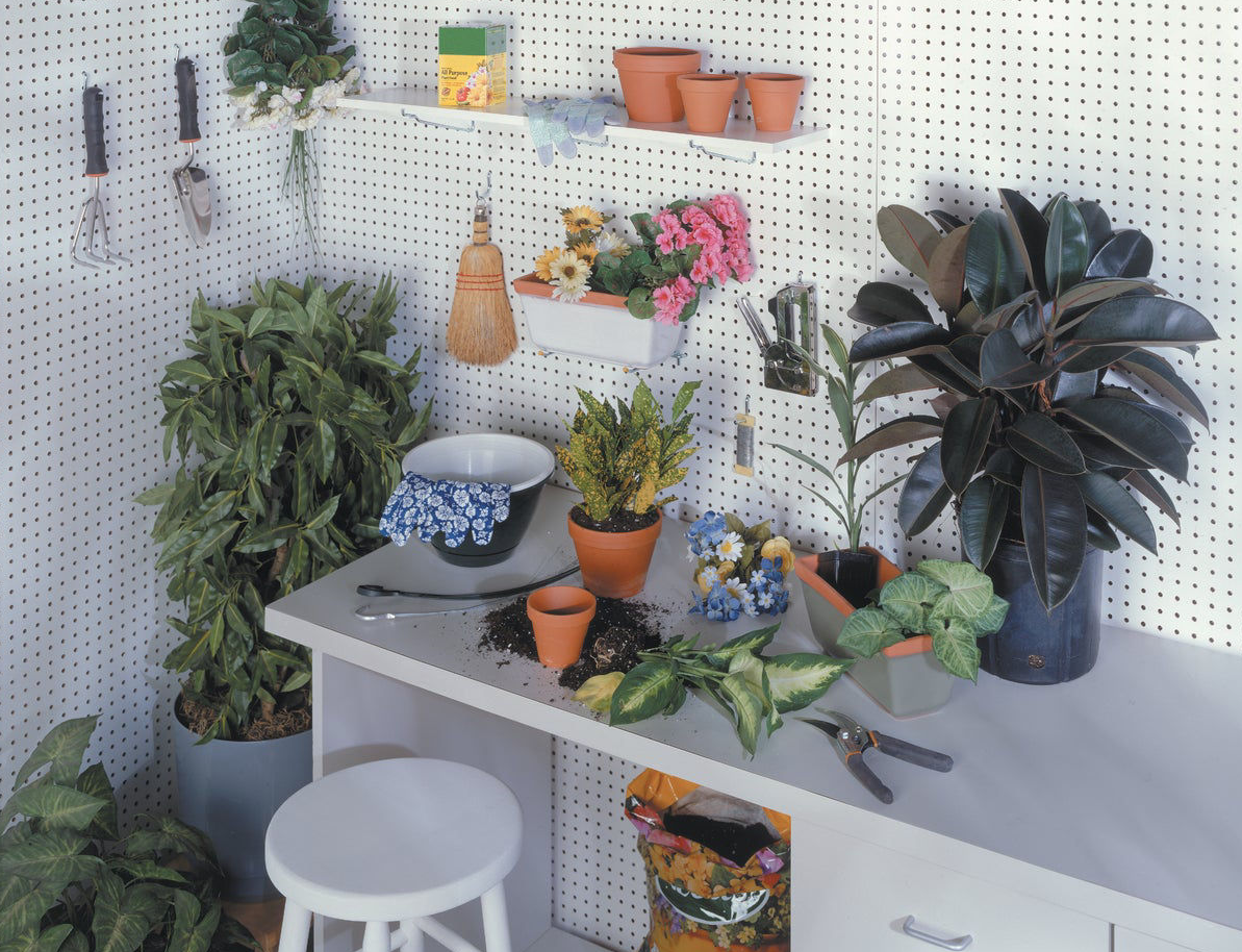 White hardboard pegboard wall organizing gardening tools and pots above white potting bench with plants and soil for home improvement.